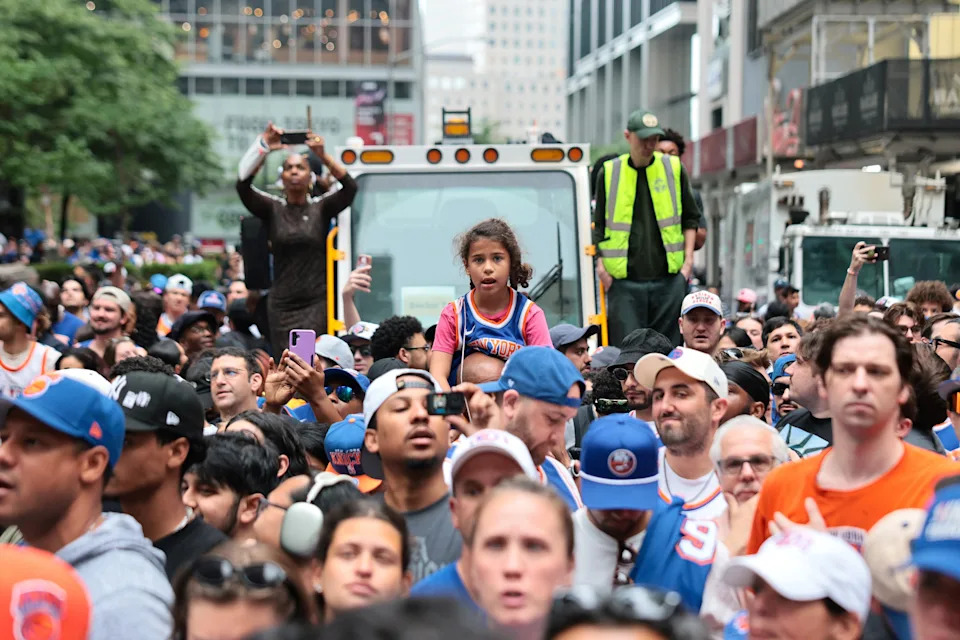 A young girl sits on a man's shoulders in a bustling city crowd; many wear sports jerseys, capturing public attention at a street event