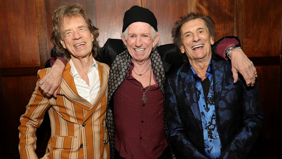 Mick Jagger, Keith Richards, and Ronnie Wood smiling together, wearing stylish jackets.
