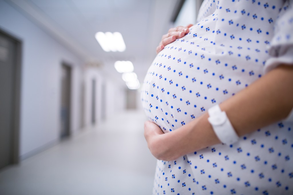 Mid-section of a pregnant woman in a hospital gown holding her belly.