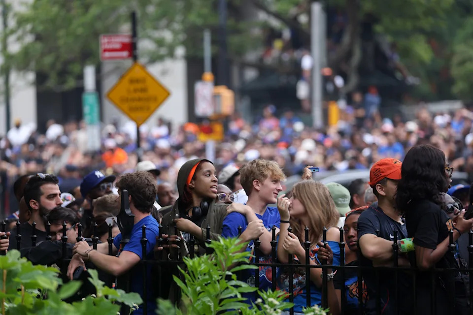 Crowd of people gathered behind a barrier, many looking eager and holding phones, suggesting a public event or street gathering