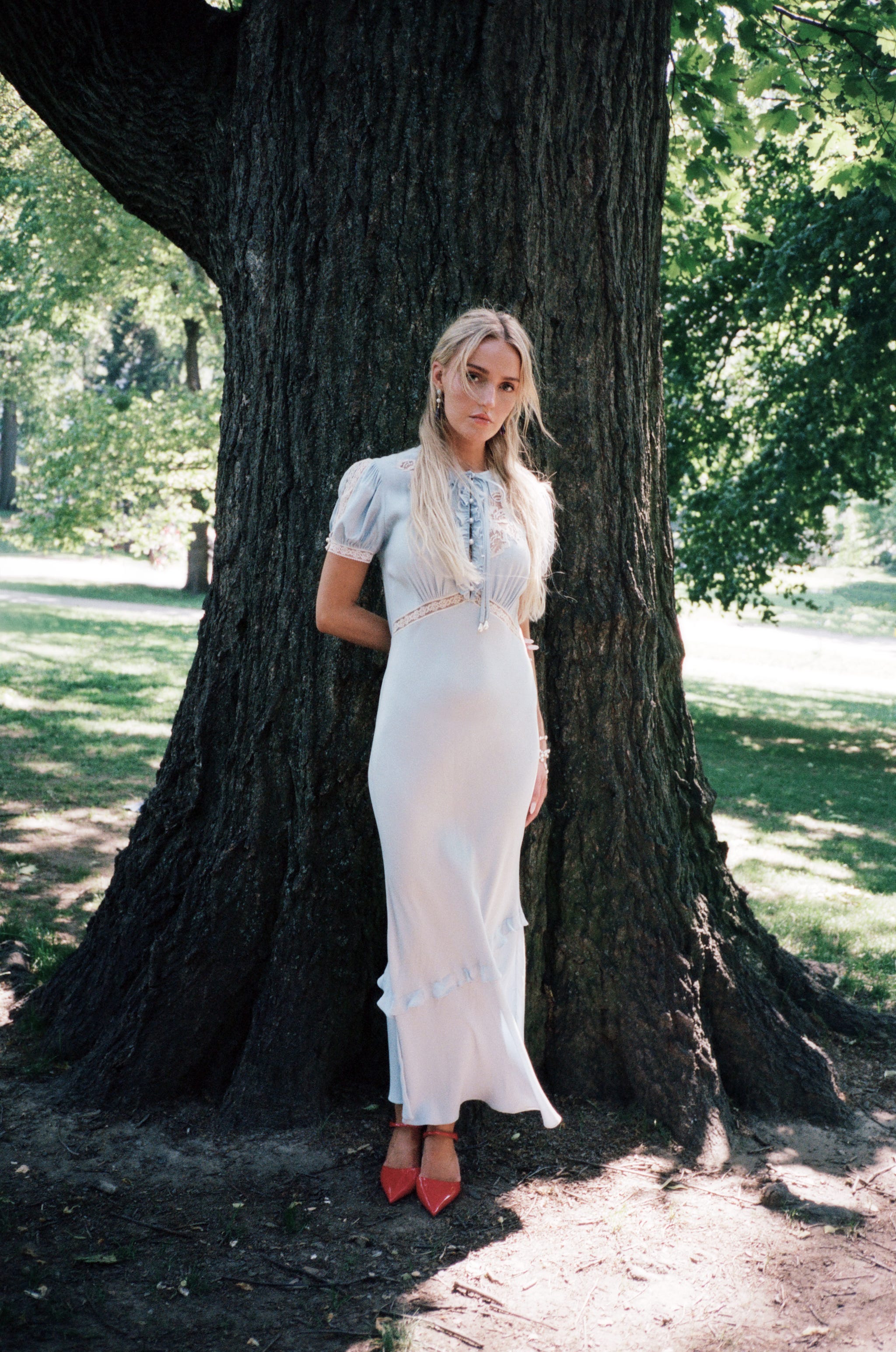 Person in a long dress with red shoes leaning against a tree in a park.