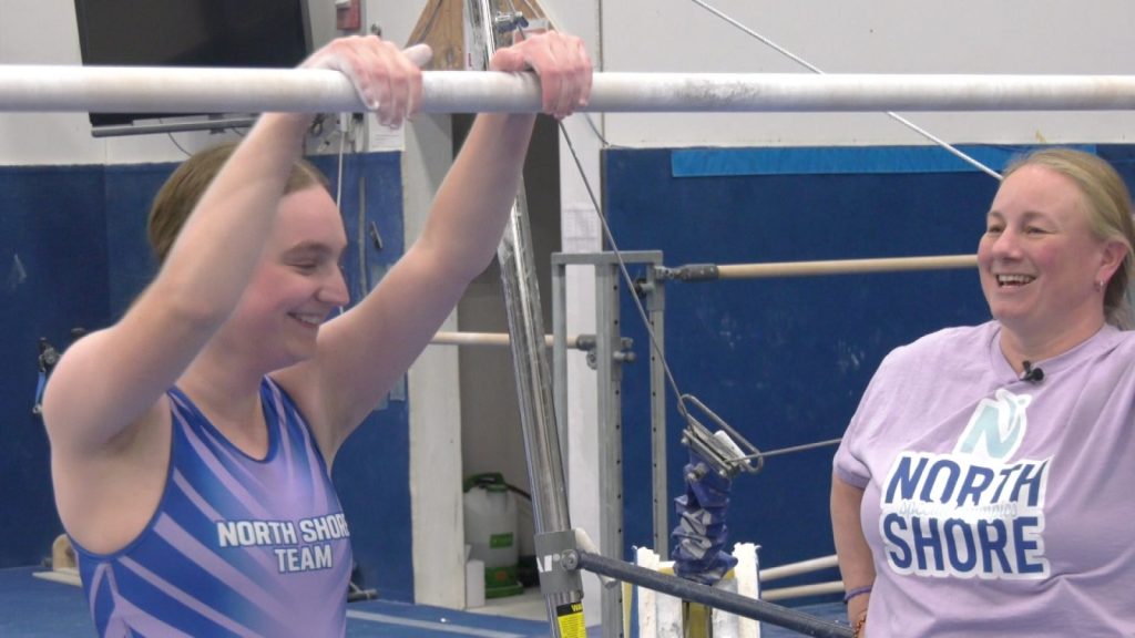 A girl hanging on a gymnastics bar in a leotard next to her coach