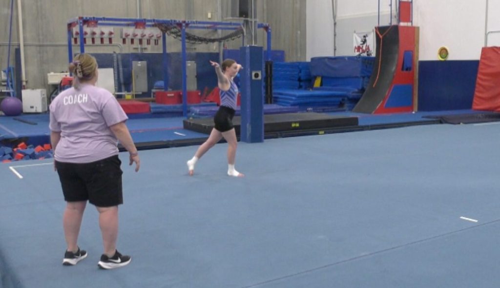 coach watching a gymnast in a purple uniform practice a floor routine