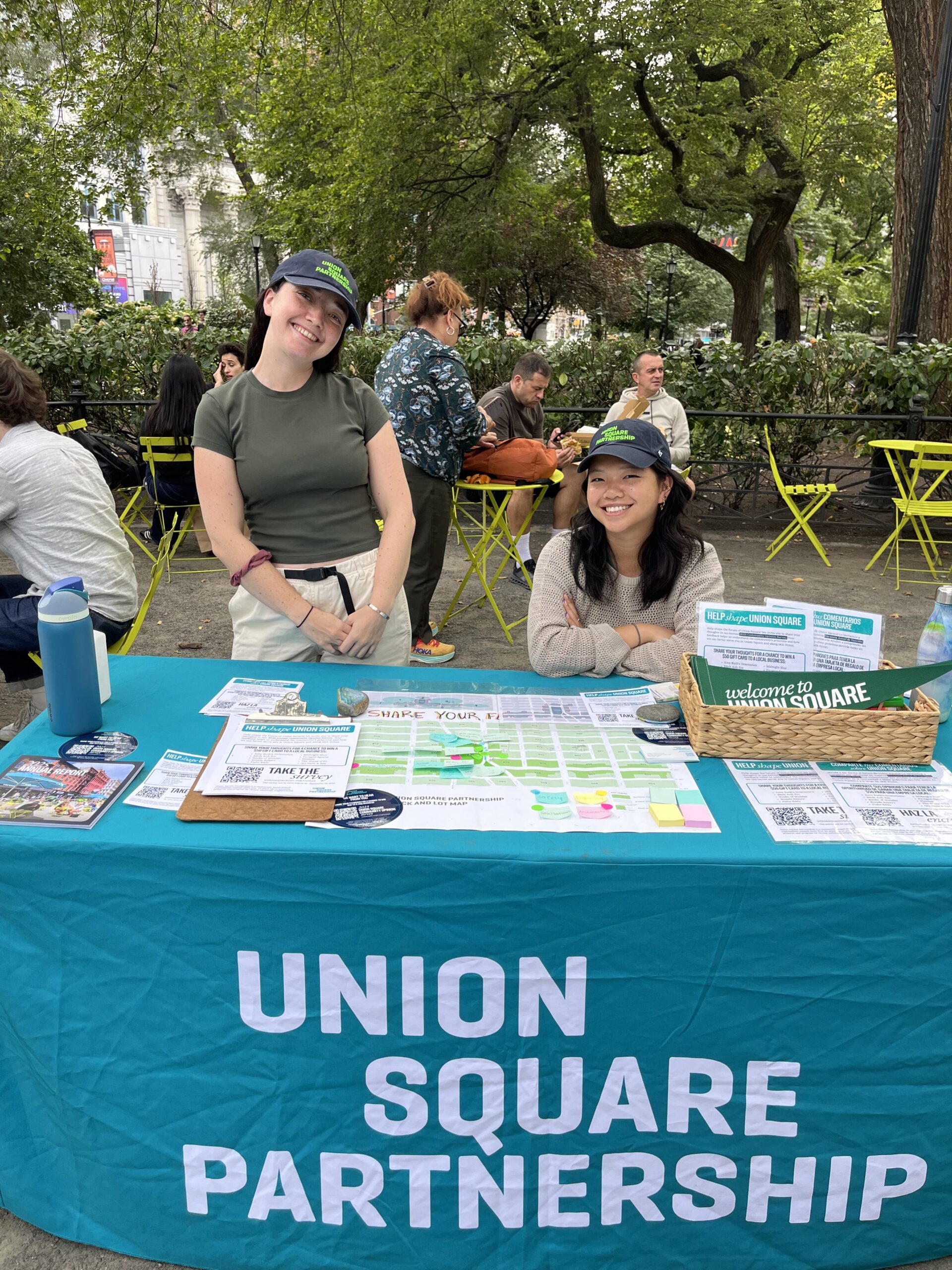 Two women stand behind a table adorned with a blue tablecloth that reads "UNION SQUARE PARTNERSHIP." They are smiling at the camera, wearing matching hats. The table displays pamphlets, a map, sticky notes, and a water bottle. In the background, people are seated at green chairs, surrounded by trees and greenery, creating a lively park atmosphere.