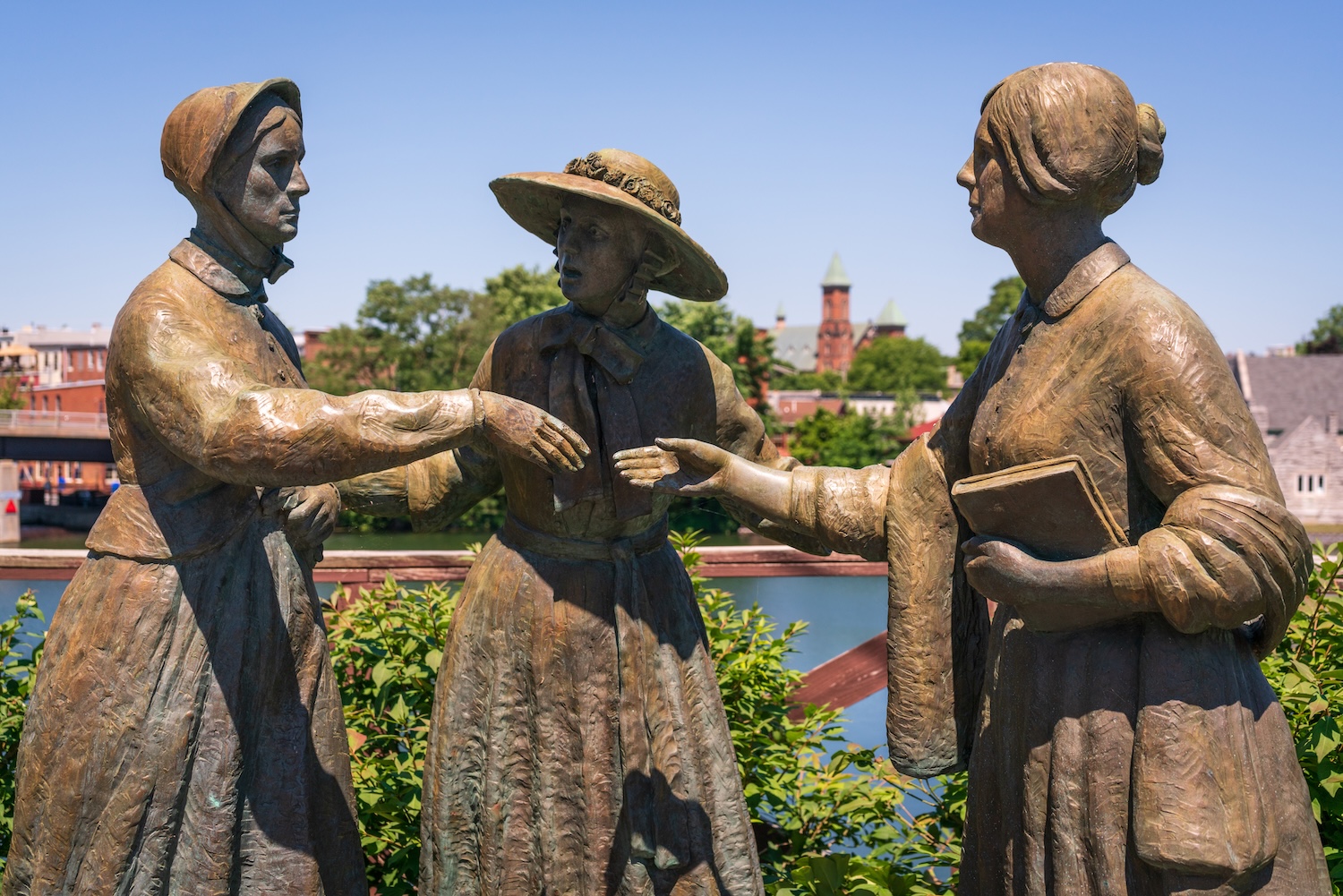 Bronze statues of three suffrage-era figures gathered in conversation beside a riverfront.