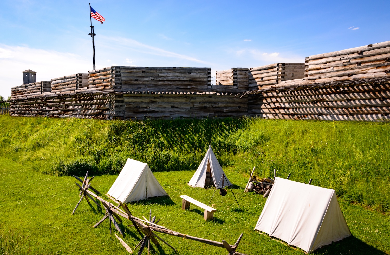Reconstructed wooden fort with canvas tents and an American flag on a grassy field.