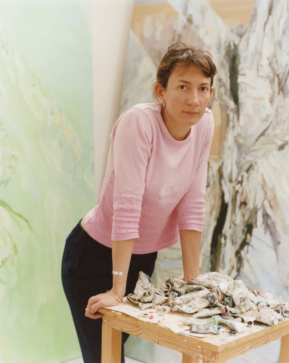 A woman in a pink long-sleeved shirt leans over a paint-splattered wooden table piled with crumpled paint tubes, with large abstract paintings visible behind her in the studio.