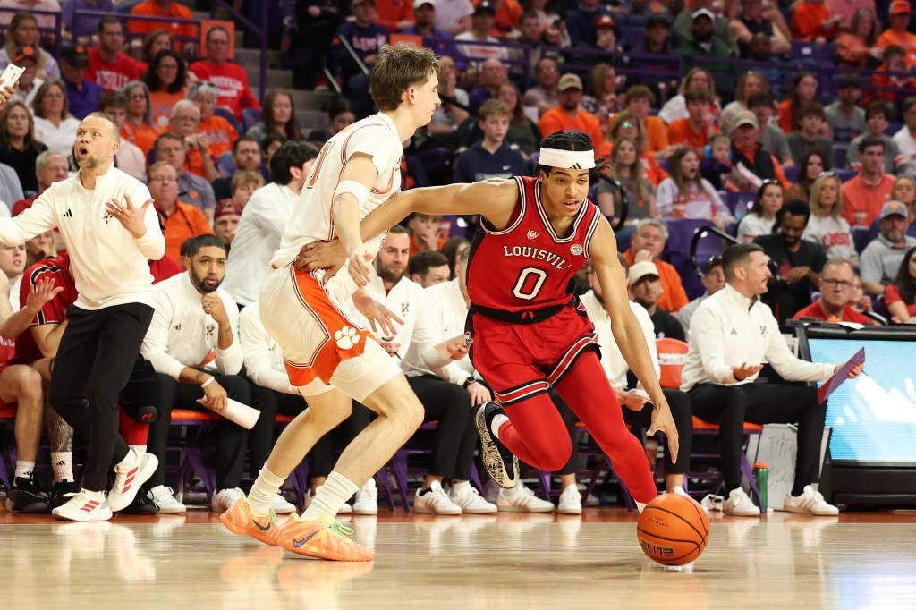 A Louisville player dribbles the ball past a Clemson player as a coach gestures from the sidelines.