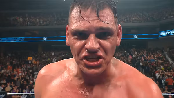 A close-up of Gunther, a wrestler in WWE, appears intense and focused, with sweat on his face and a determined expression. The audience can be seen in the background, creating an energetic atmosphere.