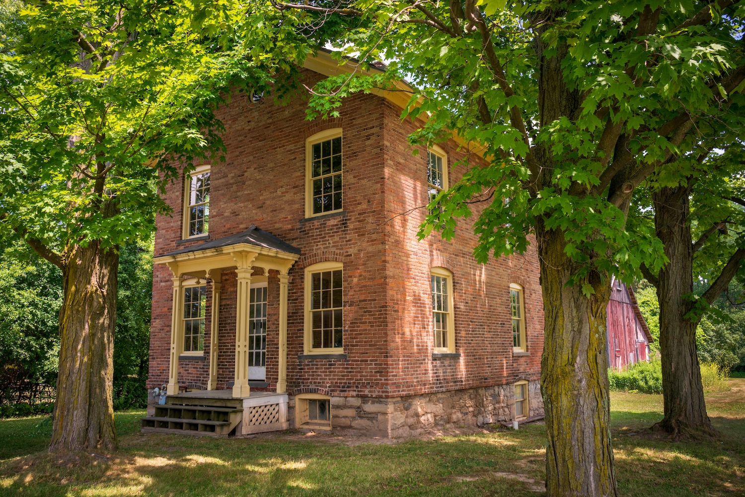 Historic brick farmhouse surrounded by mature maple trees, with a small front porch and shaded lawn.