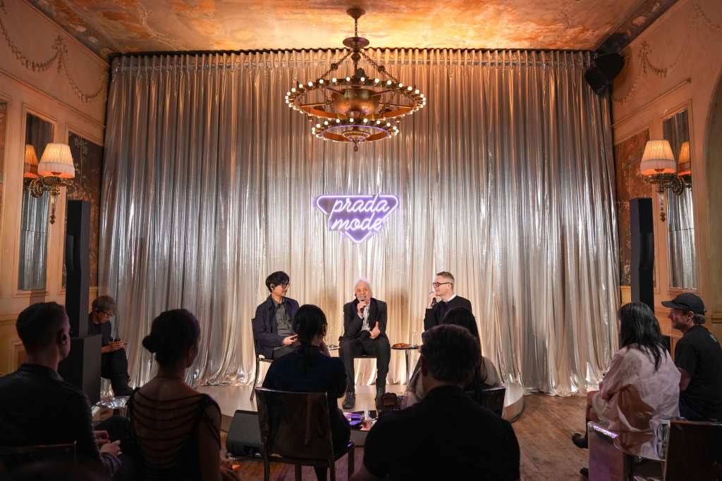 Hideo Kojima, Abel Ferrara, Nicolas Winding Refn sitting in a row on a stage in front of a large silver curtain and a purple neon sign that says "Prada Mode."