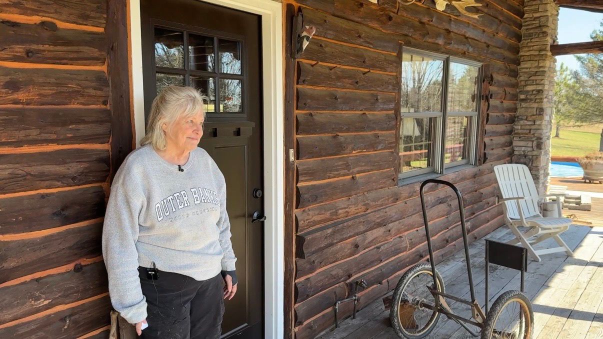 Diane Sattler stands outside of the log cabin she helped build by hand. (Spectrum News/Natalie Mooney)