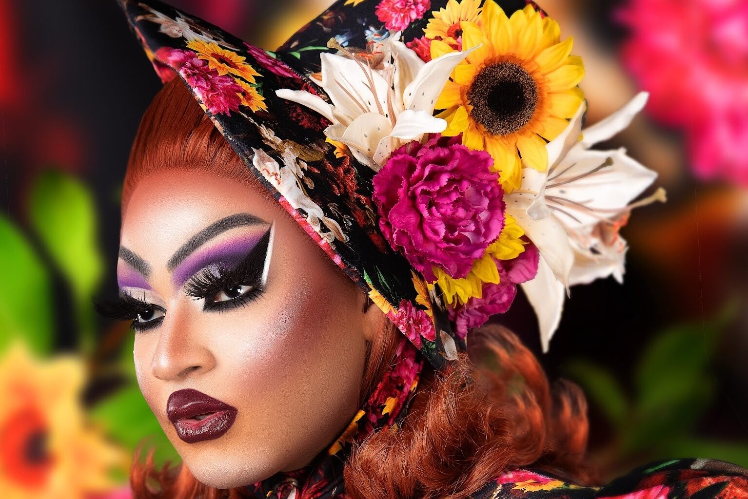 close-up side profile photo of a drag queen in heavy makeup wearing a hat decorated with artificial flowers
