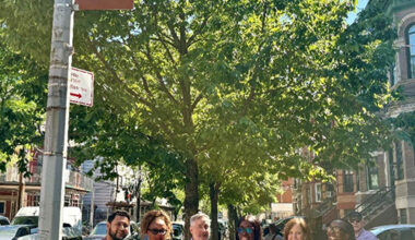 Smiling people standing next to a pole with red sign attached at the top and historic row homes in the background