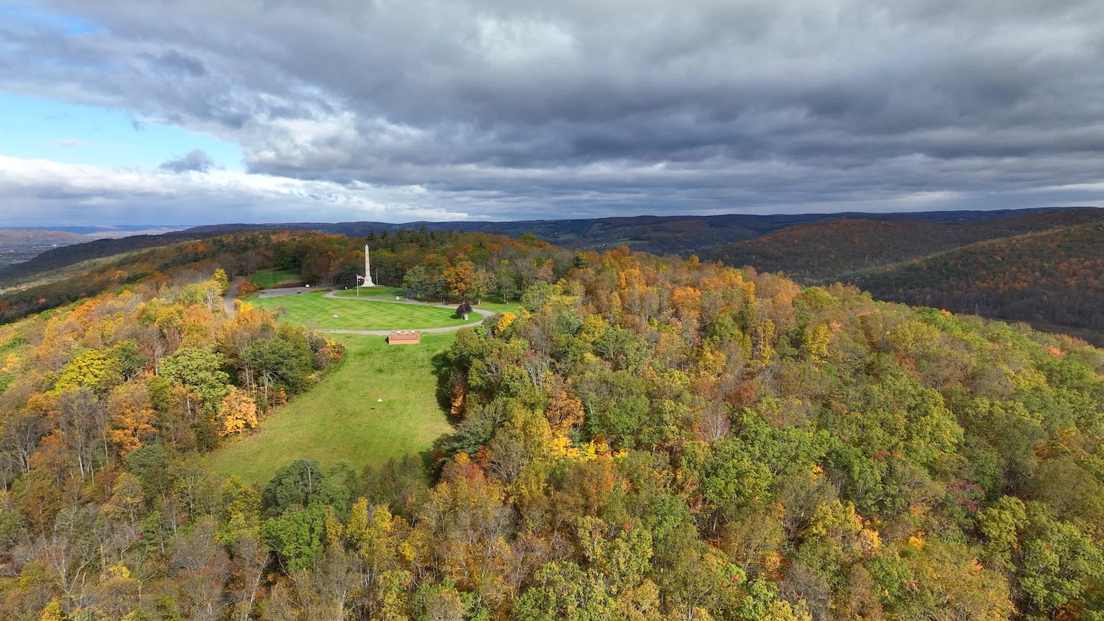 Aerial view of forested hills in autumn, with a monument and open meadow beneath dramatic clouds.