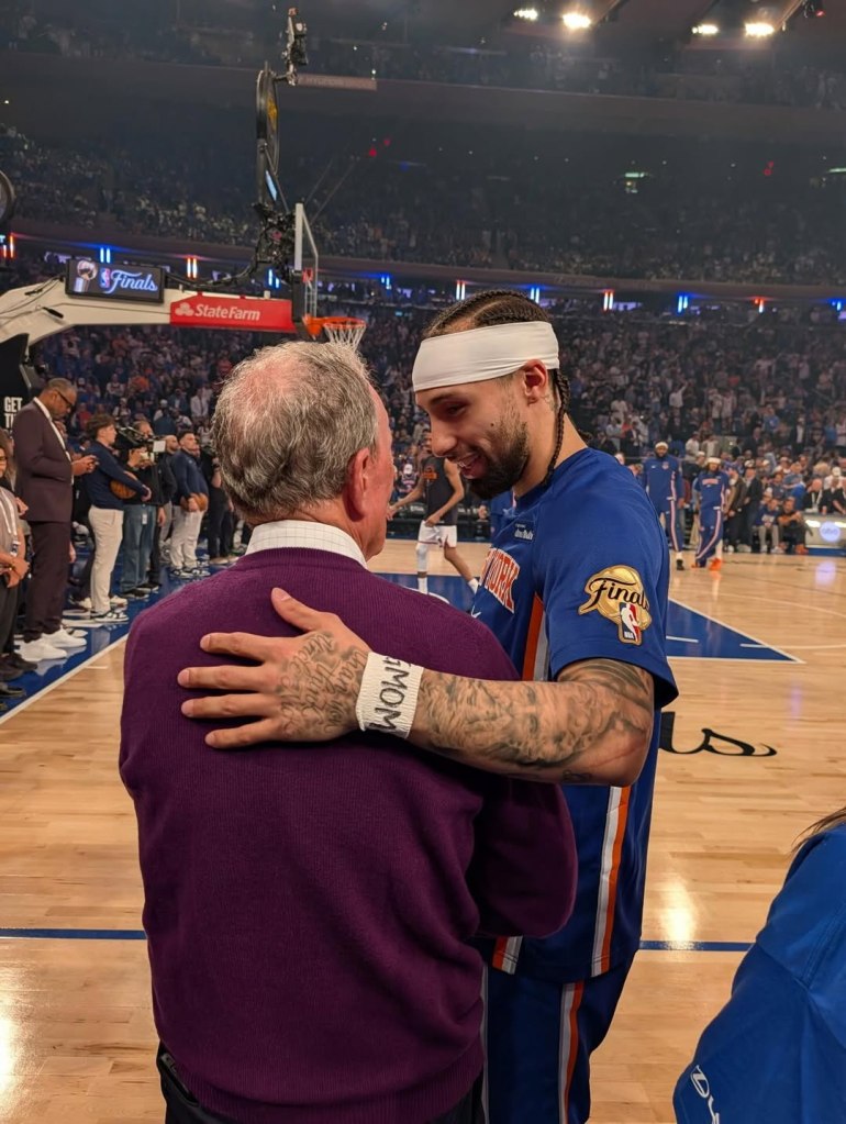 A basketball player in a blue jersey with a white headband, and an older man in a purple sweater, embracing on a basketball court.