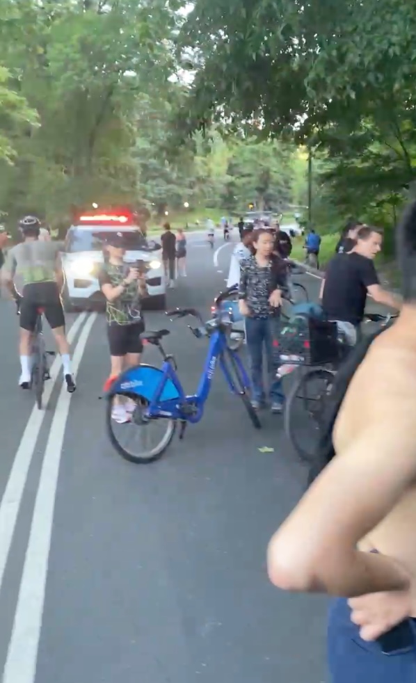Cyclists and a police car with flashing lights on a road.