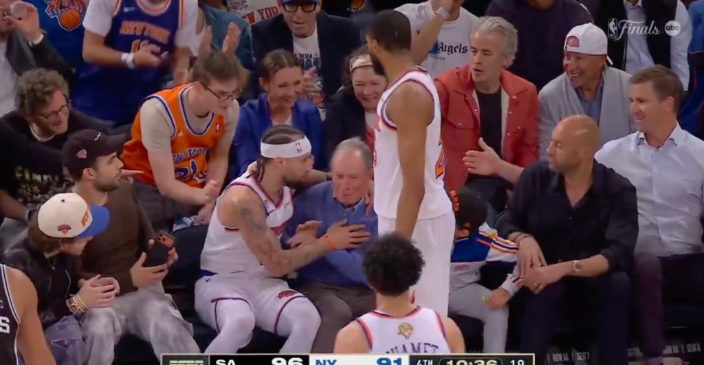 A basketball player in a white uniform with a headband reaches into the stands to grab an older man who has fallen forward.