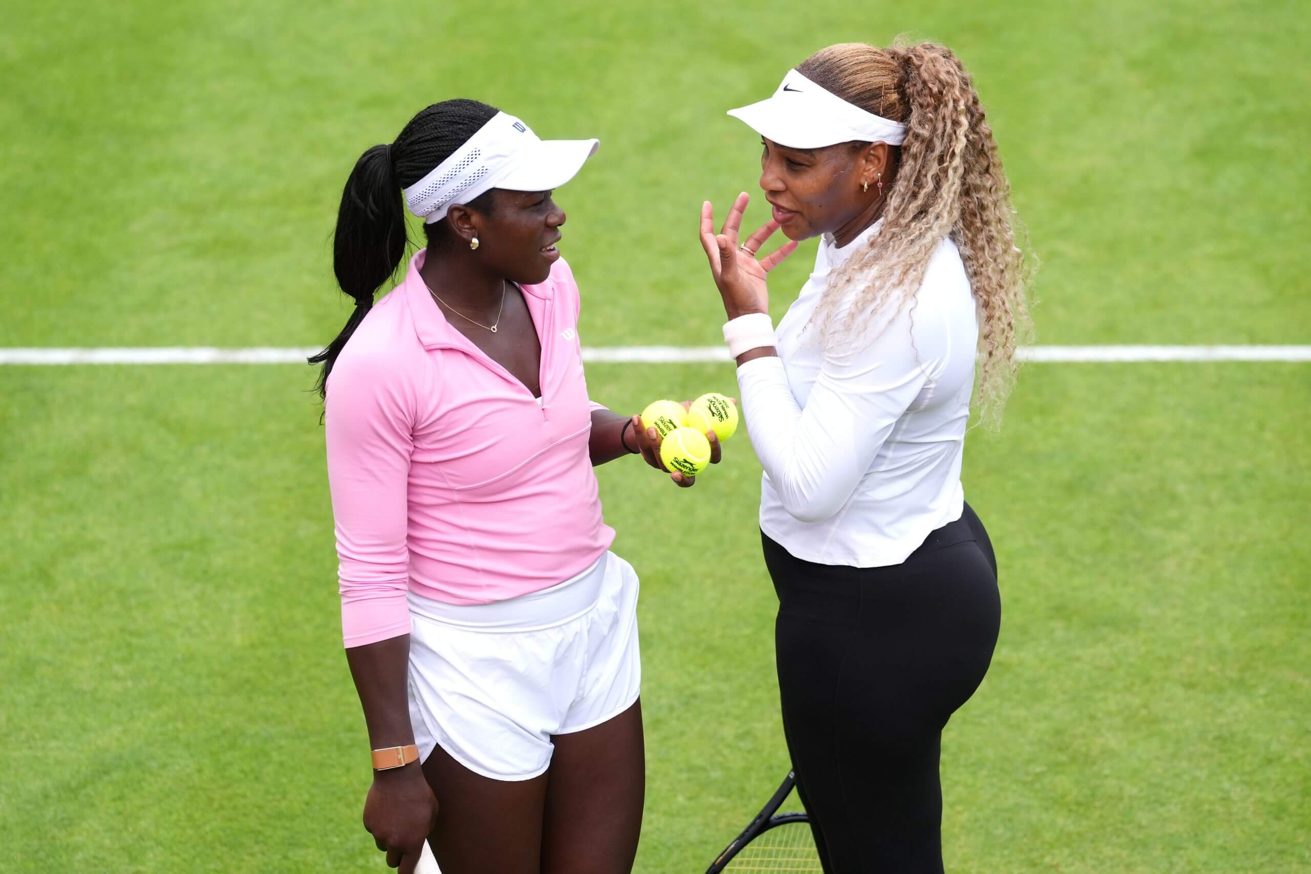 Victoria Mboko (left) and Serena Williams (right) talk on a grass tennis court, with Mboko holding three tennis balls.