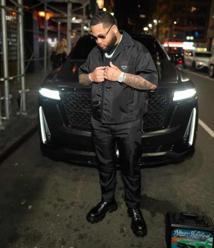 A man in a black shirt, pants, and boots stands in front of a dark car on a city street at night.