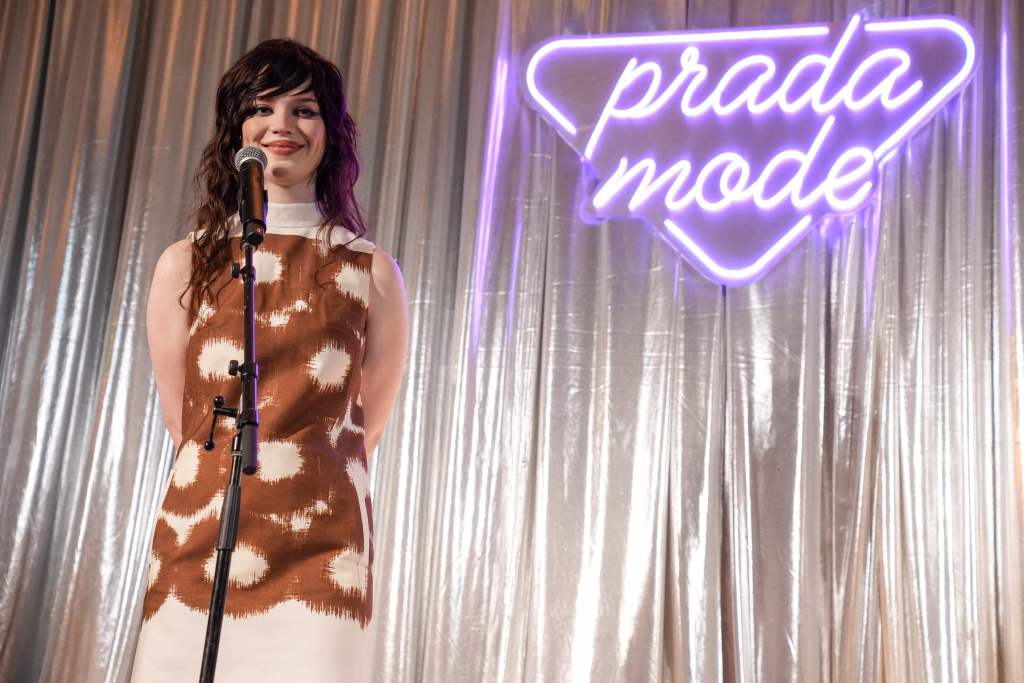 Sophie Thatcher stands next to a neon says that says "Prada Mode" in purple light.