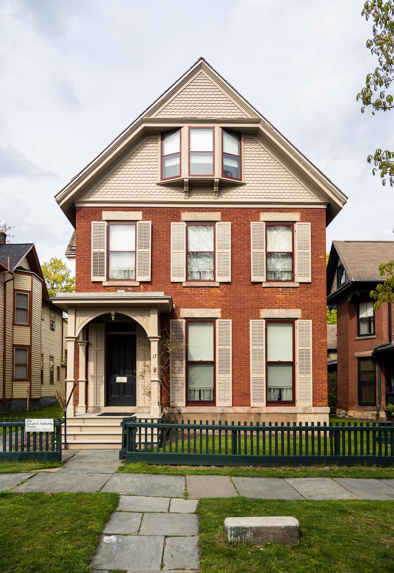 Three-story red-brick historic house with cream shutters, a gabled roof, and fenced front yard.