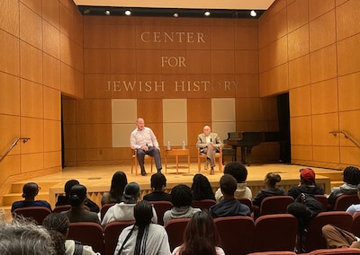 Two men sit on a stage in front of a crowd of people.