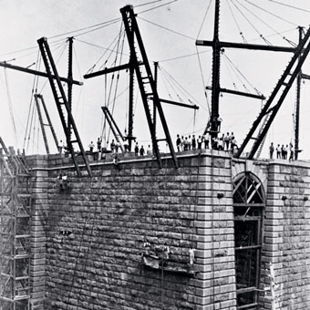 Historic black and white photograph of a large stone bridge under construction, featuring scaffolding and workers positioning large metal frameworks.