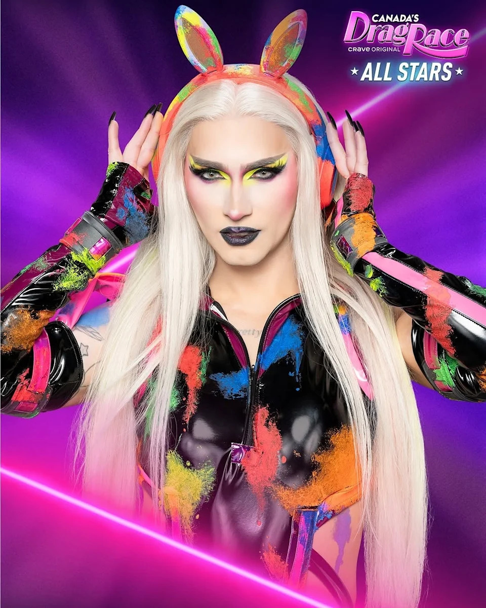 Person in bold futuristic outfit with long blonde hair, striking makeup, and colorful accessories poses confidently with hands on headphones