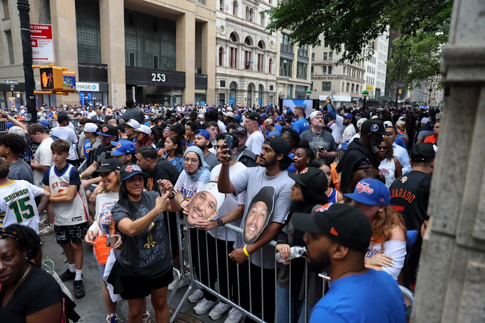A crowd of people gather on a city street, some holding masks and signs, likely at a public event or rally