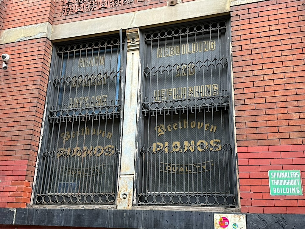 Vintage storefront featuring large, decorative iron gates with the text 'Beethoven Pianos' and details about rebuilding and refinishing pianos, set against a brick wall.
