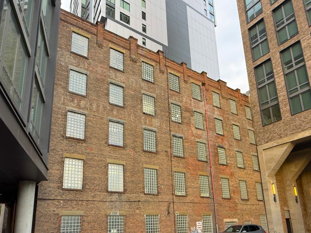 View of a brick building with glass block windows, flanked by modern structures in an urban environment.