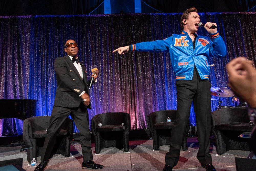 Two performers on stage, one in a tuxedo and sunglasses, the other in a blue jacket, both holding microphones.