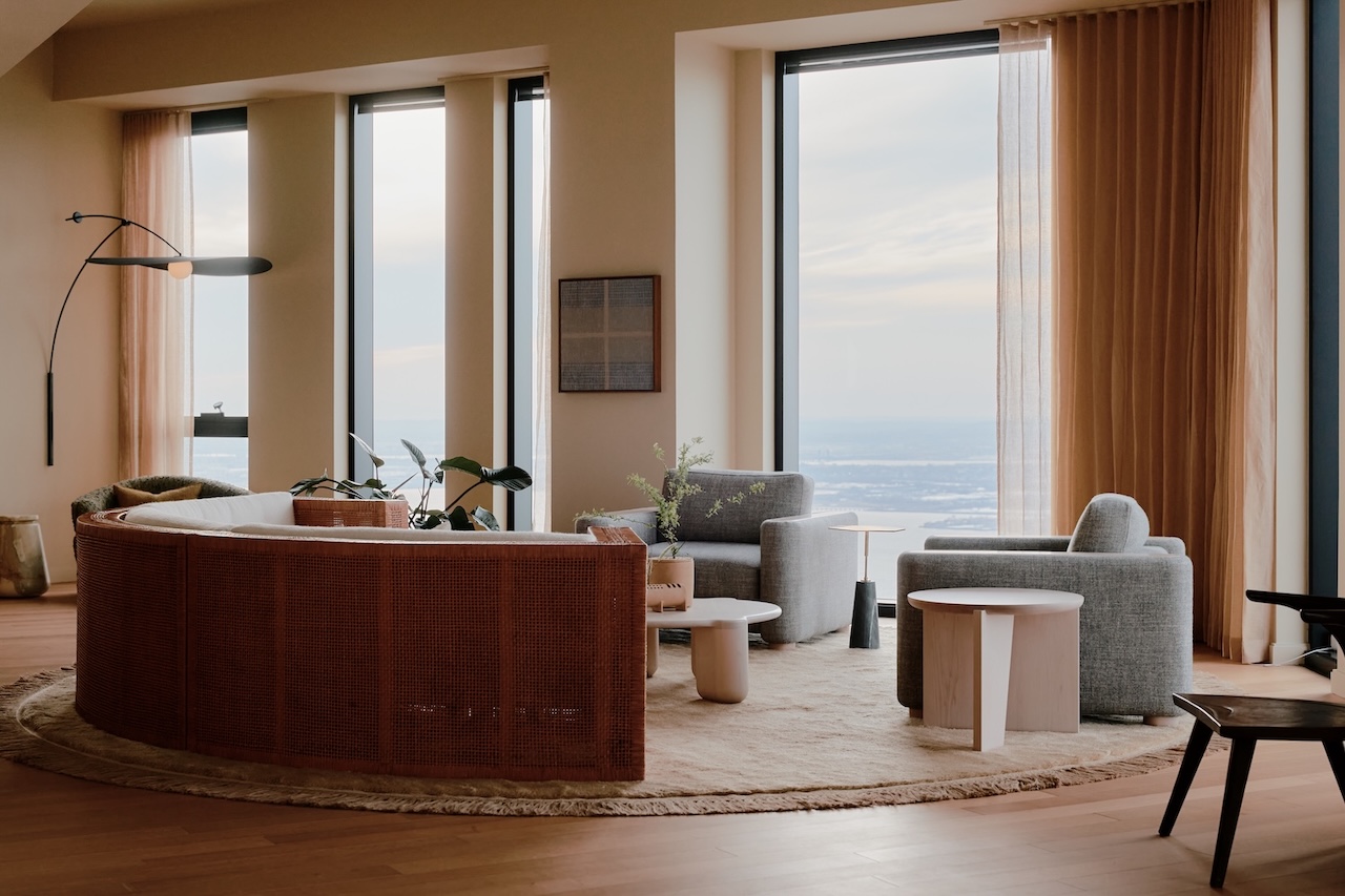 Modern living room with large windows, curved sofa, two armchairs, round rug, coffee tables, indoor plants, and neutral-toned decor. Natural light fills the space.