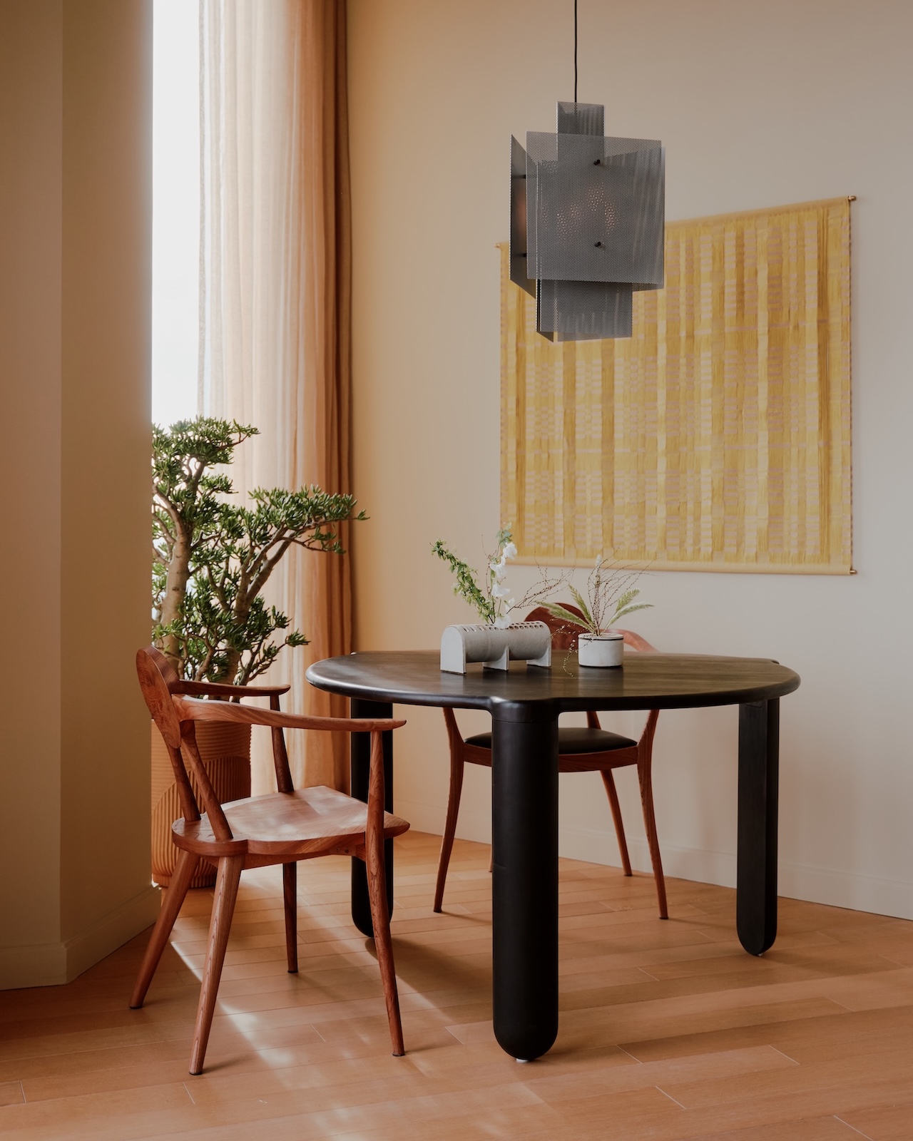 A round black table with two wooden chairs, two small plants, and minimalist decor sits near a large window with sheer curtains and a yellow wall hanging.
