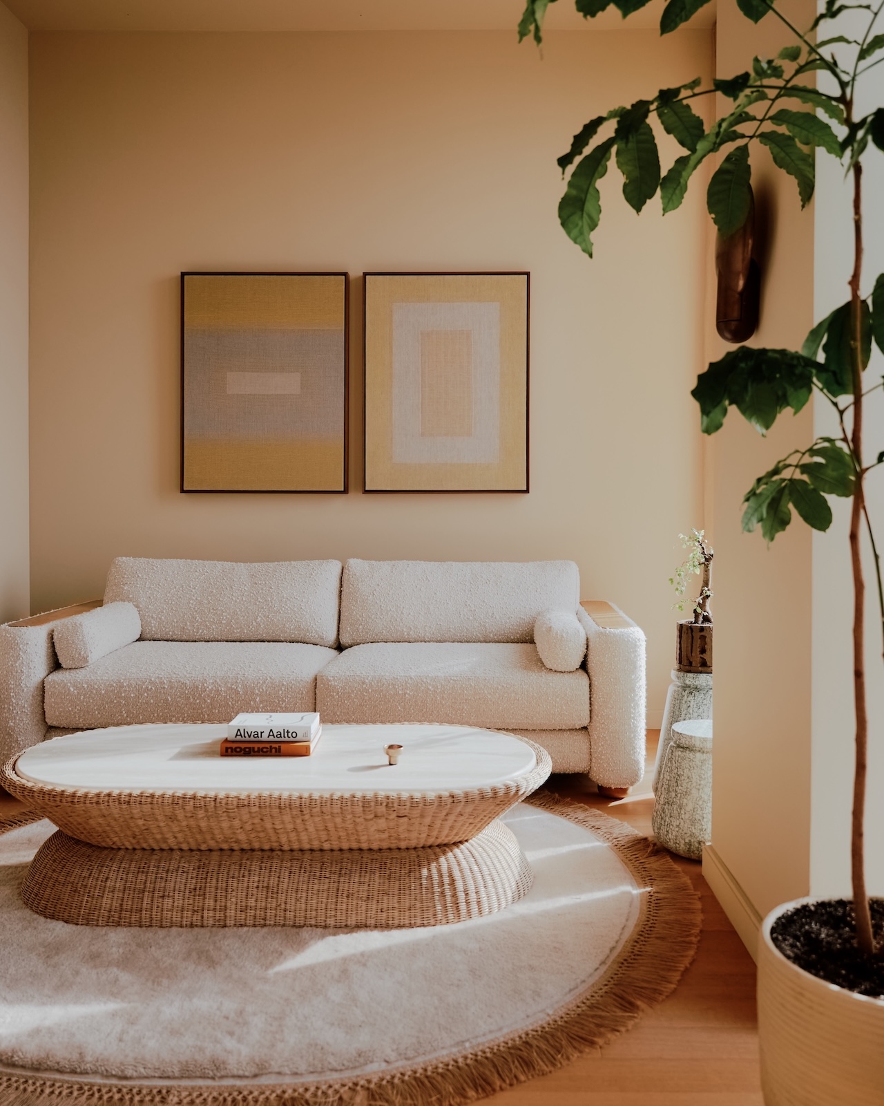 A minimalist living room with a beige sofa, two abstract wall art pieces, a round woven coffee table, and a large potted plant by the window.