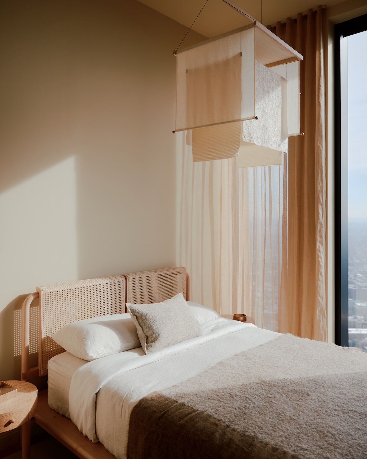 A minimalist bedroom with a wooden bed, white bedding, beige blanket, sheer curtains, and a modern hanging light fixture next to a large window with a city view.
