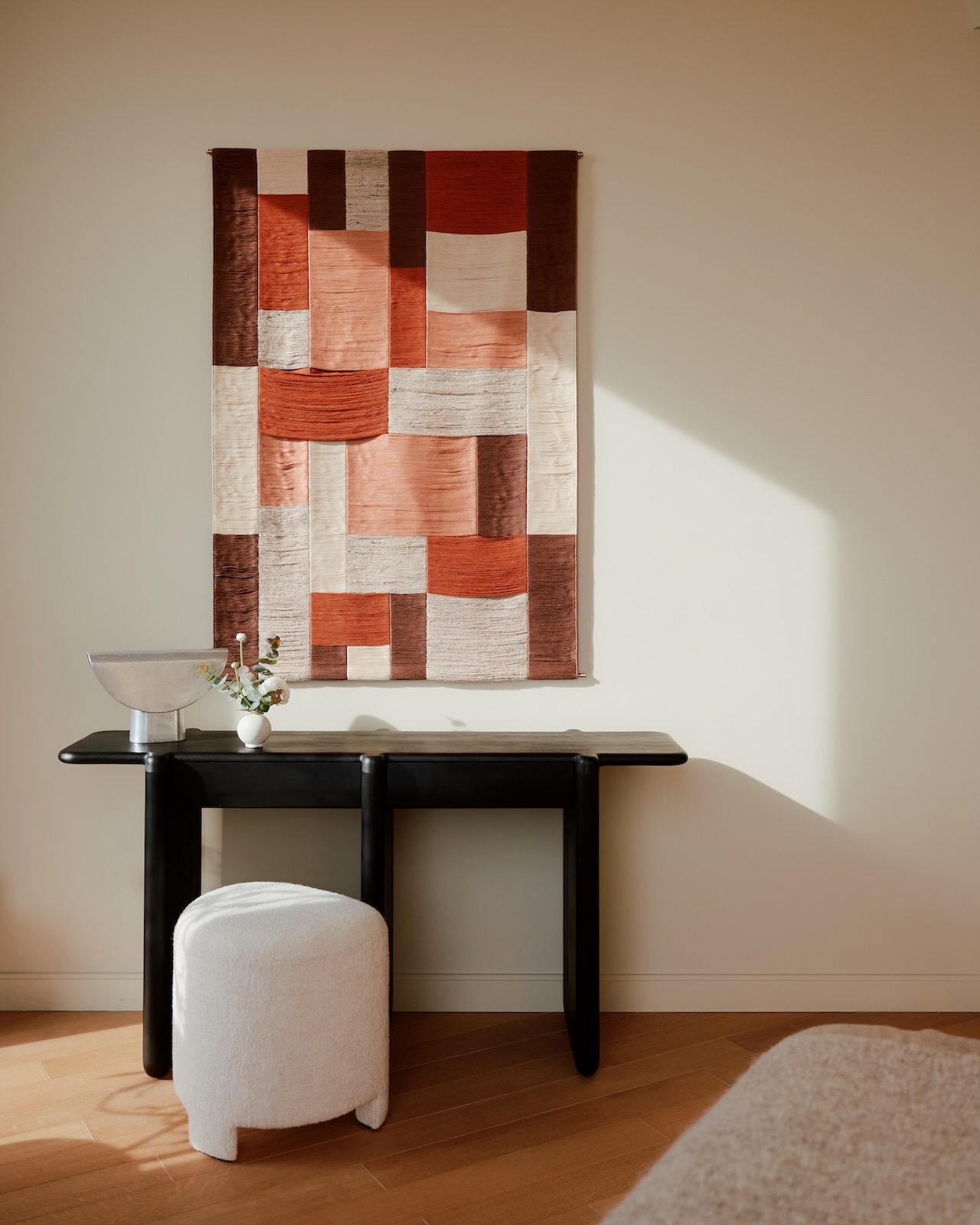 A geometric textile wall hanging in red, cream, and brown tones above a black console table with a silver bowl, small vase, and a white round stool.