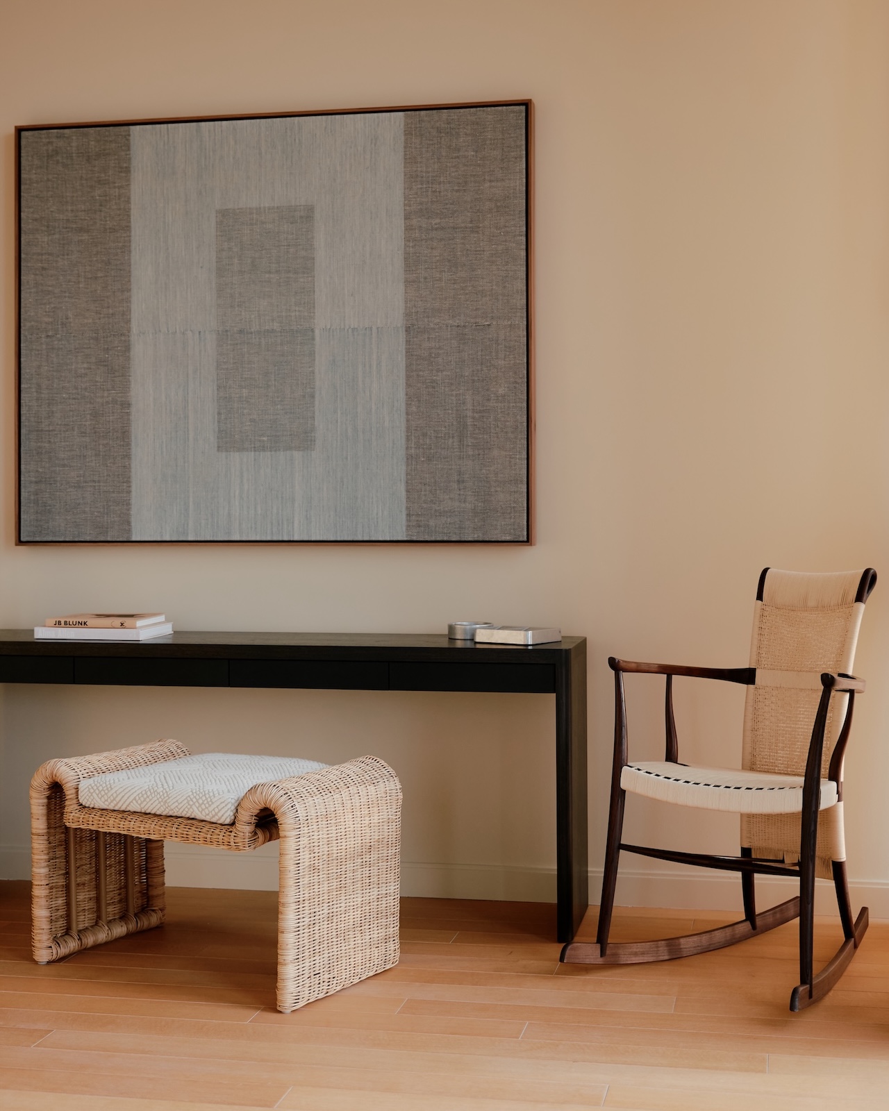 A minimalist room with a black desk, wicker stool, beige rocking chair, and large abstract artwork on a light-colored wall.