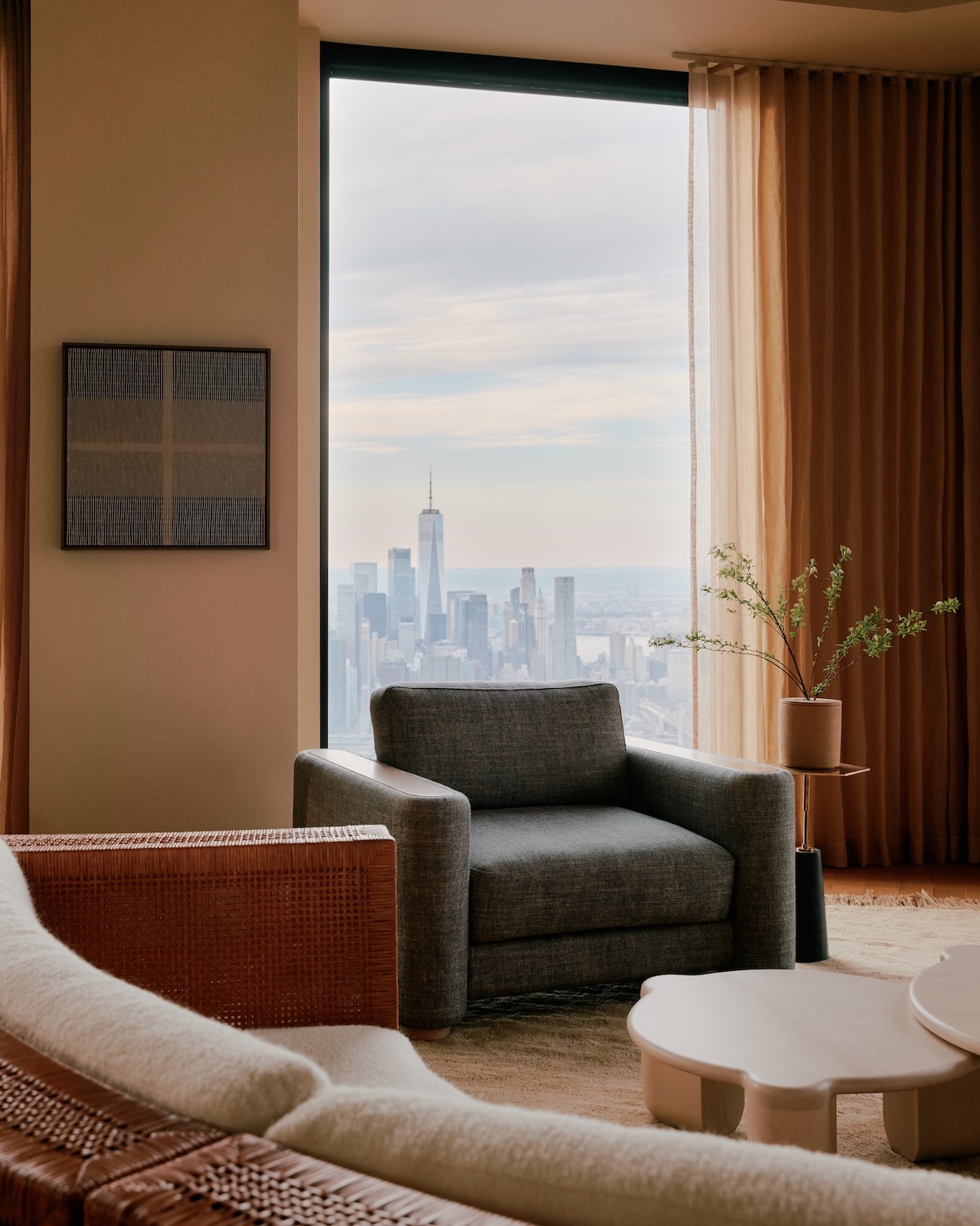 A modern living room with a gray armchair and a large window overlooking a city skyline, including a tall skyscraper, under a cloudy sky.