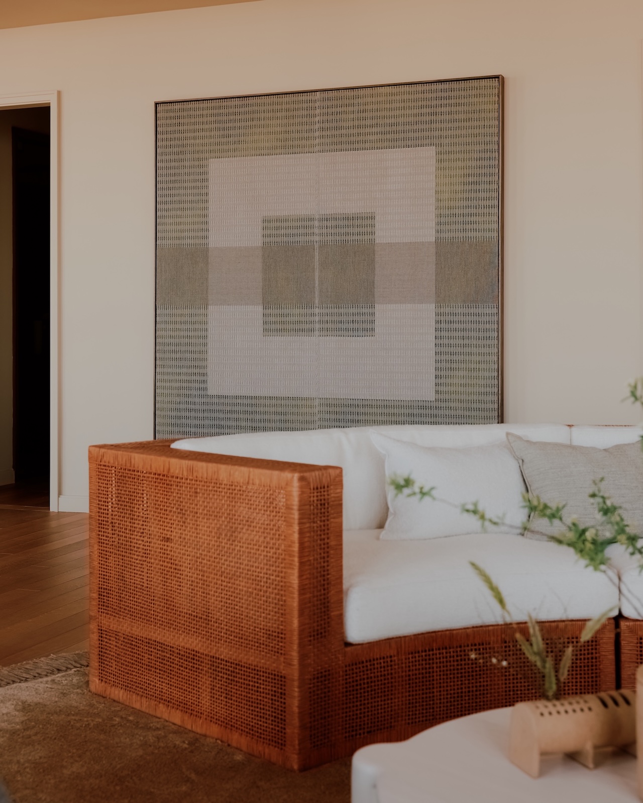 A modern living room with a rattan sofa, white cushions, a large geometric wall art, wooden floor, and a small table with a plant.