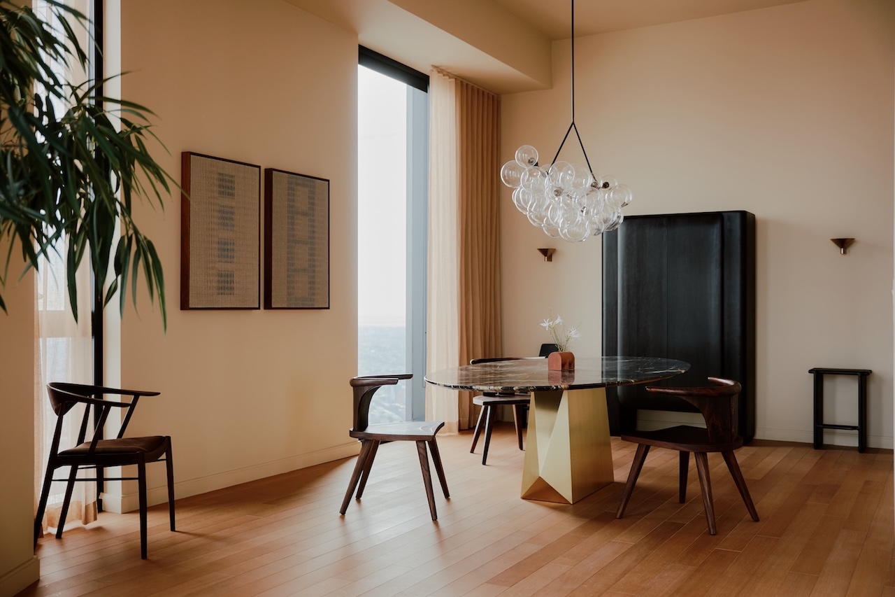 Modern dining room with a round marble table, four wooden chairs, a geometric gold base, contemporary bubble chandelier, two framed artworks, and a tall black cabinet.