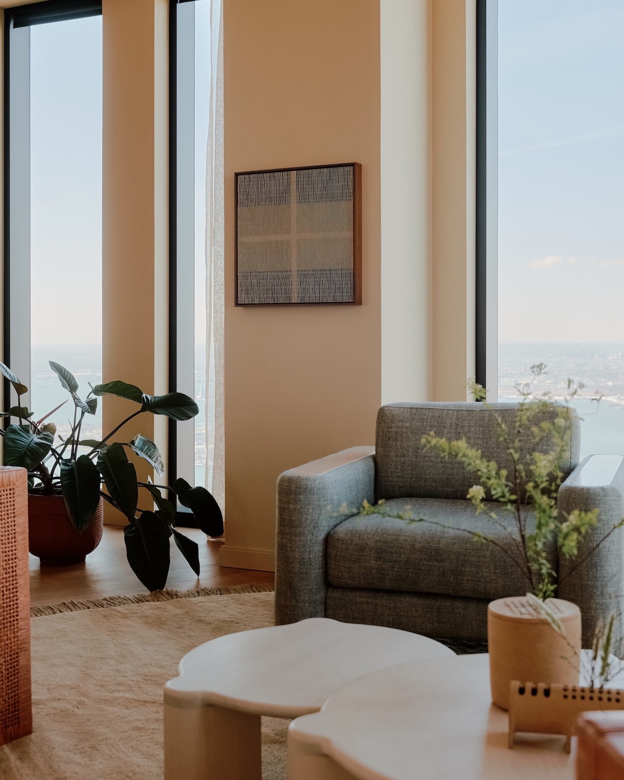 A modern living room with a gray armchair, light wood tables, potted plants, and large windows overlooking a city and water view.