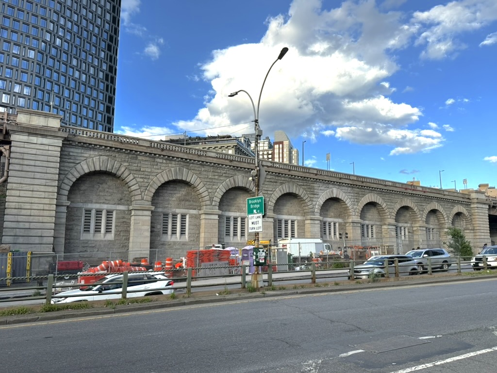 A stone archway structure along a roadway, with a modern building in the background and construction equipment visible in the foreground under a partly cloudy sky.