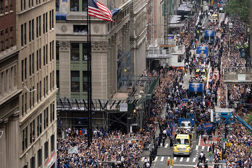 Crowds line a busy city street during a parade, with floats and vehicles moving through, while an American flag flutters overhead