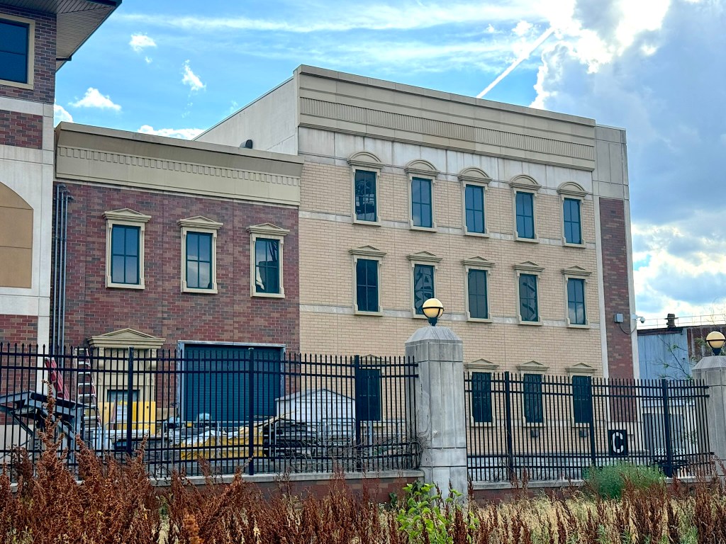 A brick building featuring a mix of architectural styles, with large windows and decorative trim. The lower level has a red brick facade, while the upper levels display a lighter color. Surrounding the building is a black fence with light fixtures.