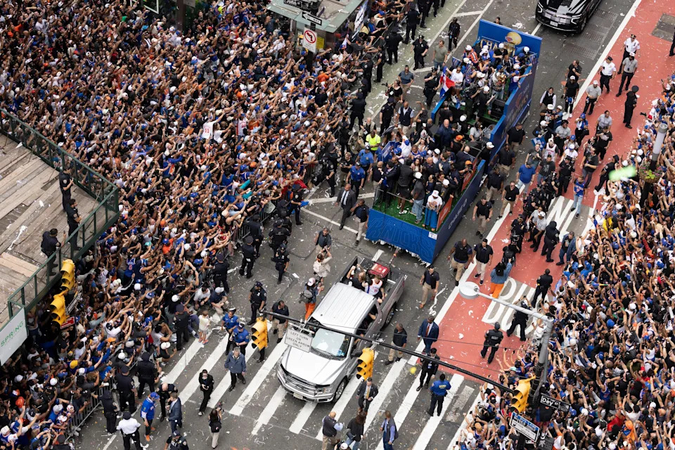 A large crowd gathers at a street parade, cheering and waving as vehicles and people pass through, surrounded by security and media personnel
