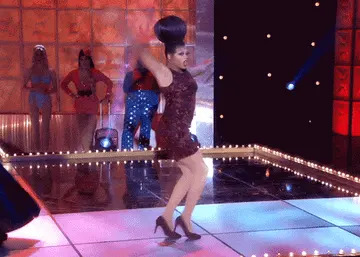 Performer in a fitted dress and elaborate headpiece dances energetically on a stage, with other performers in the background