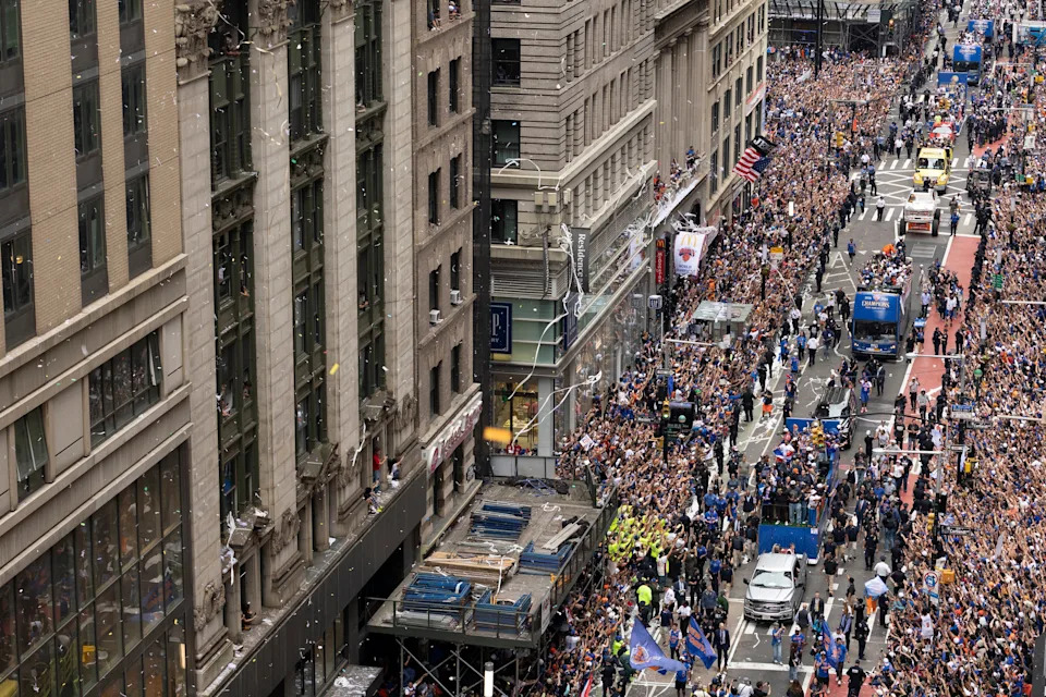 A large crowd fills a city street celebrating with a parade. Aerial view shows buildings, vehicles, and people waving flags