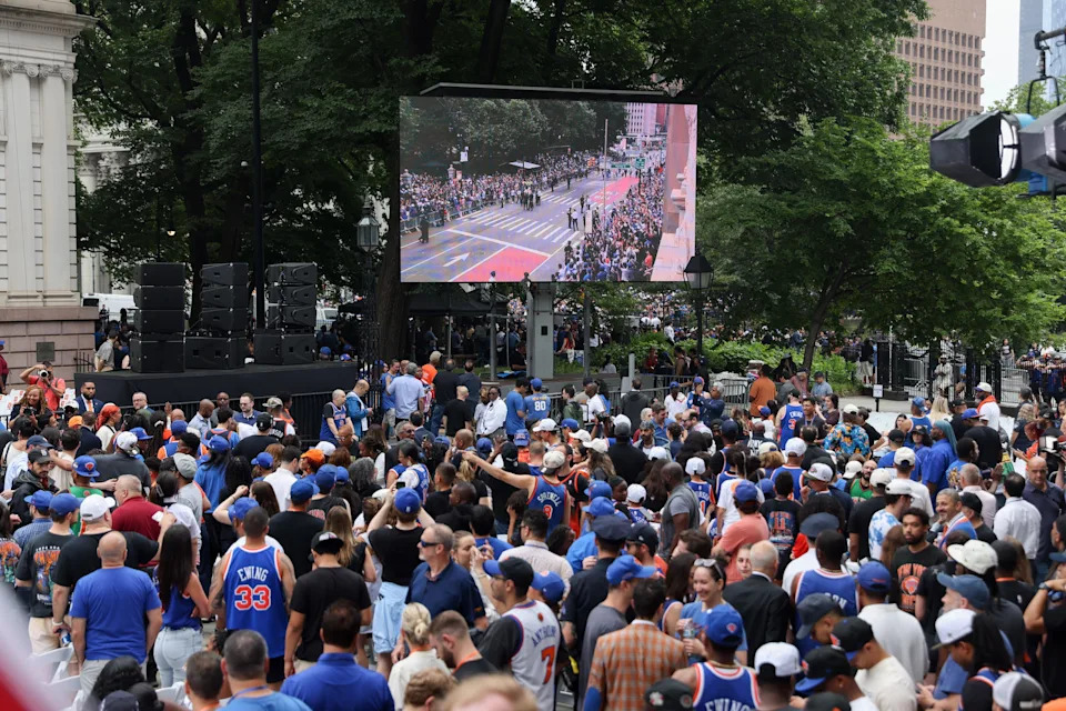 A large crowd gathers outdoors, watching a sports event on a big screen. Many are wearing sports jerseys and hats, engaged and excited